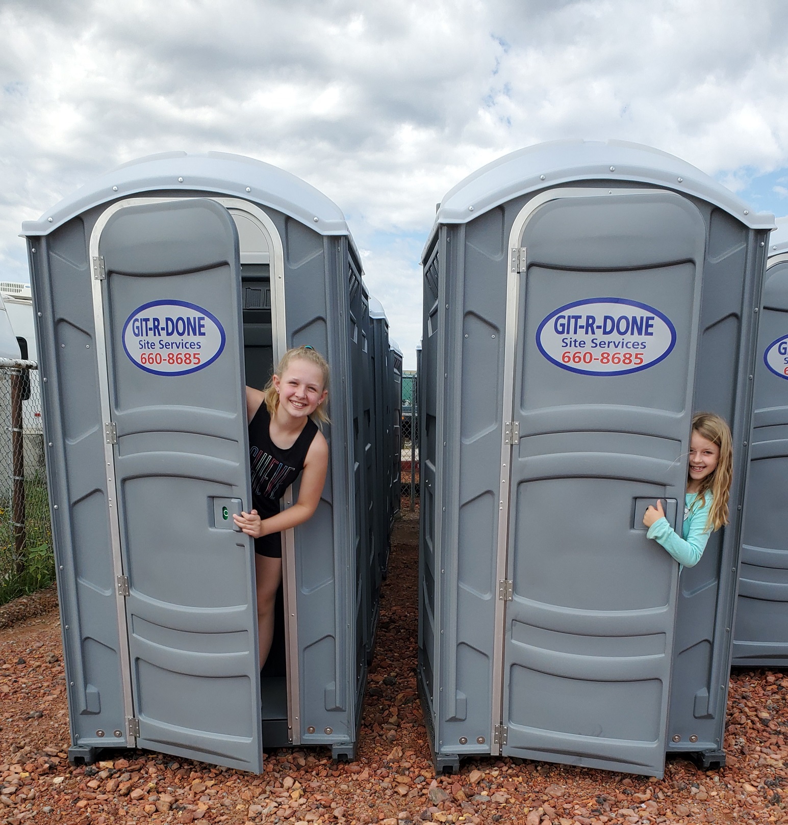 Two Portable Toilets With Two Girls Looking out and Smiling